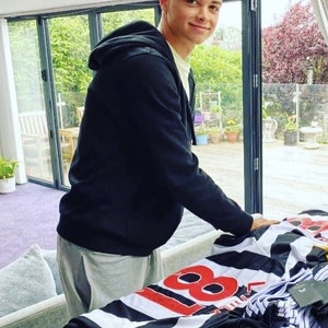 May include: A young person wearing a black hooded sweatshirt and gray sweatpants stands in front of a table with a stack of black and white striped shirts with the number 88 printed on them.