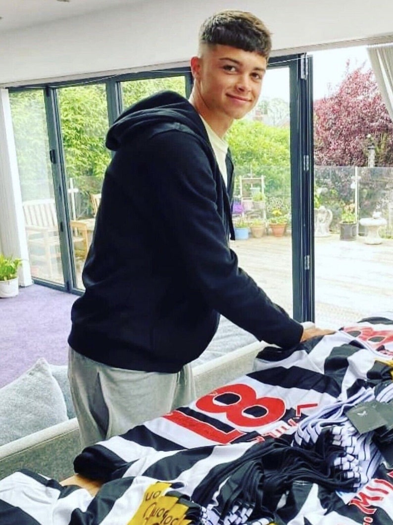 May include: A young person wearing a black hooded sweatshirt and gray sweatpants stands in front of a table with a stack of black and white striped shirts with the number 88 printed on them.