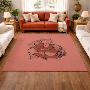 May include: A rectangular, dusty rose-colored rug featuring a black line drawing of a couple embracing, with a horse's head above them. The rug is placed in a living room with white sofas, a red sofa, and wooden side tables.