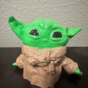 K&ouml;nnte beinhalten: Eine gr&uuml;ne und braune Figur eines Charakters aus dem Star Wars-Franchise. Der Charakter hat gro&szlig;e Ohren und gro&szlig;e Augen. Die Figur ist aus einem strukturierten Material gefertigt.