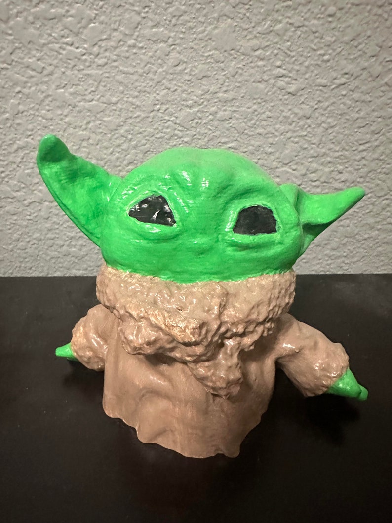 K&ouml;nnte beinhalten: Eine gr&uuml;ne und braune Figur eines Charakters aus dem Star Wars-Franchise. Der Charakter hat gro&szlig;e Ohren und gro&szlig;e Augen. Die Figur ist aus einem strukturierten Material gefertigt.