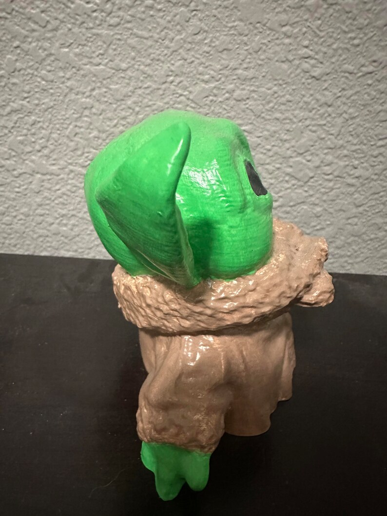K&ouml;nnte beinhalten: Eine gr&uuml;ne und braune Figur eines Charakters aus dem Star Wars-Franchise. Der Charakter hat gro&szlig;e Ohren und tr&auml;gt einen braunen Mantel.