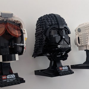 May include: Three LEGO Star Wars helmets: Luke Skywalker (Red Five) in white, red, and black; Darth Vader in black; and a Scout Trooper in white. Each helmet is mounted on a black stand with a nameplate.