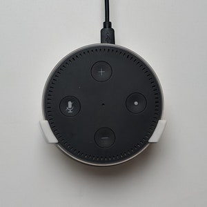 May include: A black circular smart speaker with four buttons, a microphone icon, and a white wall mount. The speaker is plugged in with a black cord.