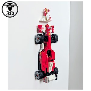 May include: A red and white toy race car with black wheels and a small figurine on top. The car is mounted on a white wall. The image includes the text "PRINT IT 3D" in a circular logo.