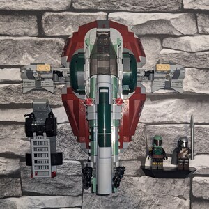 May include: A Lego Star Wars set featuring a green and red spaceship with two minifigures. The spaceship has a cockpit, wings, and a tail. The minifigures are a Mandalorian and a Boba Fett.