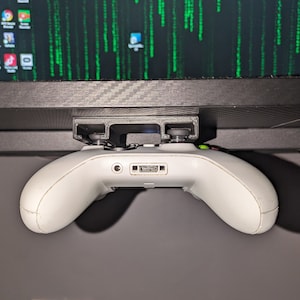 May include: A white Xbox controller is mounted to a black 3D printed bracket, which is attached to the underside of a black desk or shelf.