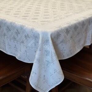 Puede incluir: Un mantel blanco con un diseño floral plateado. El mantel está colocado sobre una mesa de madera, con una esquina doblada para mostrar el patrón. La tela parece suave, posiblemente lino. Adecuado para una mesa de comedor.