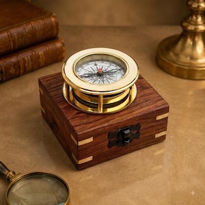 May include: A brass nautical compass with a white dial and black markings sits atop a square dark wood box. The box features brass corner inlays and a black metal latch on the front. The compass has a circular frame and a glass top cover.