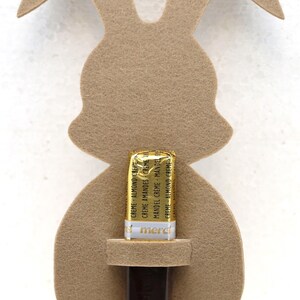 May include: A brown felt bunny cutout with a slot for a chocolate bar. The chocolate bar is wrapped in gold foil and has the text "CREME ALMOND CREME" printed on it.