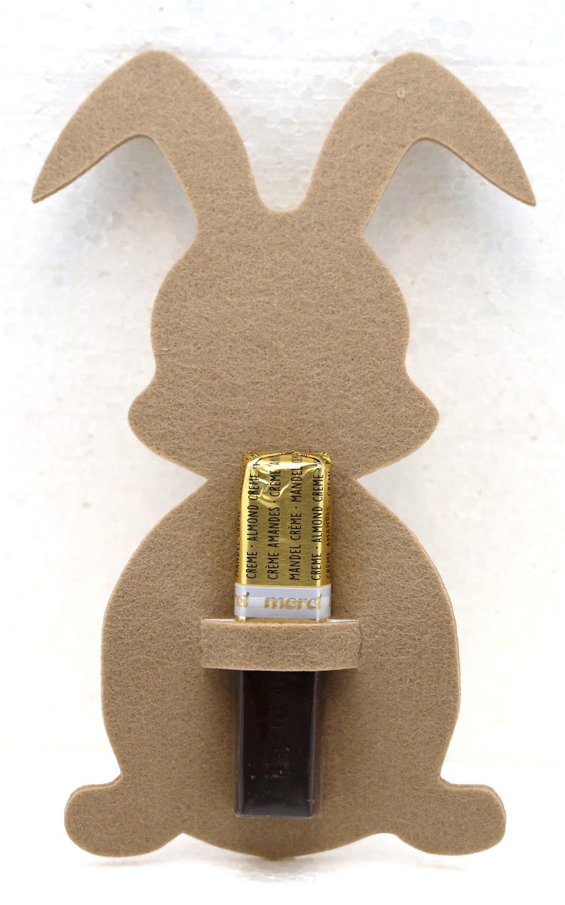 May include: A brown felt bunny cutout with a slot for a chocolate bar. The chocolate bar is wrapped in gold foil and has the text "CREME ALMOND CREME" printed on it.