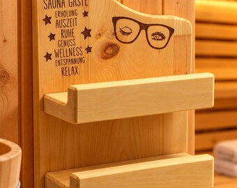 May include: A light-colored wooden wall-mounted rack with two shelves. The board features black text reading BRILLENHALTER FÜR SAUNA GÄSTE, ERHOLUNG, AUSZEIT, RUHE, GENUSS, WELLNESS, ENTSPANNUNG, RELAX, star icons, and a graphic of eyeglasses with eyes.