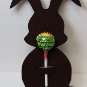 May include: A brown felt bunny cutout with a green and white lollipop in its mouth. The lollipop is a Vimto brand apple flavor.