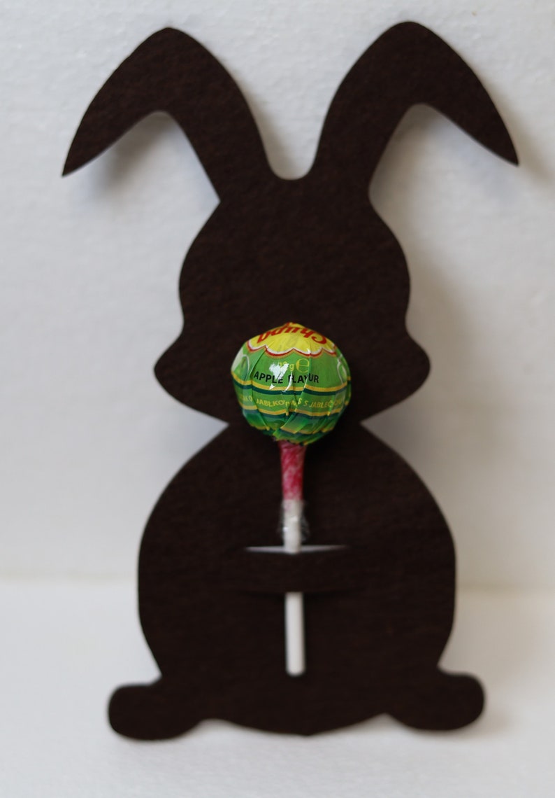 May include: A brown felt bunny cutout with a green and white lollipop in its mouth. The lollipop is a Vimto brand apple flavor.