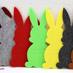 May include: Seven felt bunny shapes in different colors: red, gray, green, yellow, dark gray, brown, and a light brown. The bunnies are all the same size and shape, and they have a 2 mm felt thickness. The text "2 mm Farbauswahl" is printed below the bunnies.