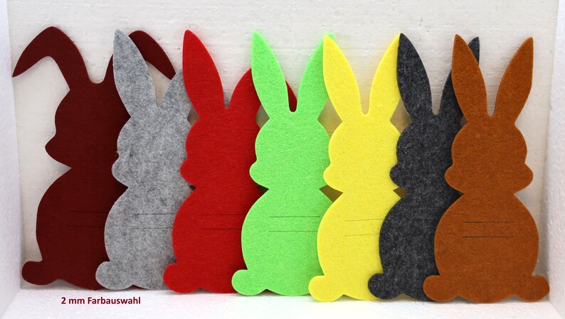 May include: Seven felt bunny shapes in different colors: red, gray, green, yellow, dark gray, brown, and a light brown. The bunnies are all the same size and shape, and they have a 2 mm felt thickness. The text "2 mm Farbauswahl" is printed below the bunnies.