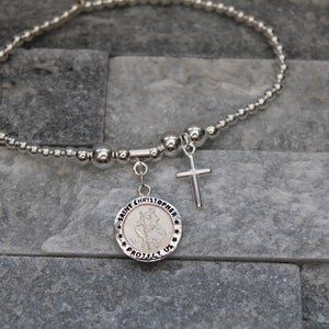 Könnte beinhalten: Ein silbernes Perlenarmband mit einem Saint-Christopher-Anhänger und einem silbernen Kreuzanhänger. Der Anhänger trägt den Text "Saint Christopher Protect Us".