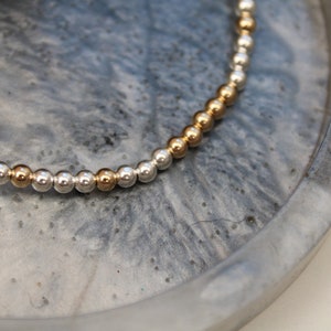 May include: A silver and gold beaded bracelet on a grey background.