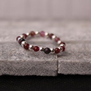 Peut inclure: Une bague ornée de petites pierres rouges foncées et de perles argentées alternées. La bague est posée sur une surface grise texturée, mettant en valeur son design et son contraste de couleurs.