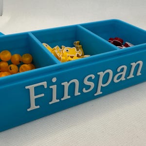 Könnte beinhalten: Ein türkisfarbener Kunststoff-Organizer mit drei Fächern, die jeweils kleine Spielsteine enthalten. Der Organizer trägt das Wort "Finspan" in weißen Buchstaben auf der Vorderseite. Ein passender türkisfarbener Deckel ist teilweise sichtbar.