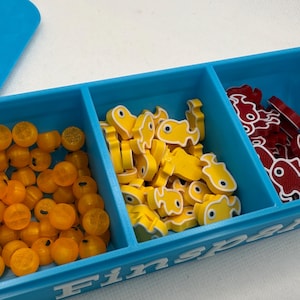 Könnte beinhalten: Ein blauer Kunststoff-Organizer mit drei Fächern, gefüllt mit Spielsteinen. Die Fächer enthalten orangefarbene runde Teile, gelbe fischförmige Teile und rote fischförmige Teile. Das Wort "Finspan" ist auf der Seite sichtbar.