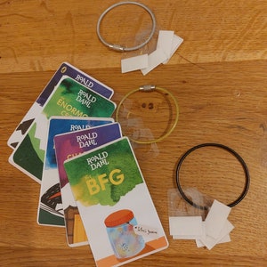 May include: A set of five illustrated cards featuring Roald Dahl books. The cards are stacked on a wooden surface. The top card features the book "The BFG" with a jar of "Fizzy Dream" on the front. The cards are surrounded by various keychains and blank labels.