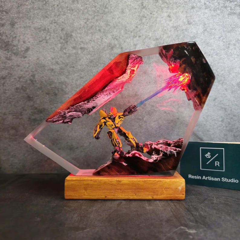 May include: A yellow and black robot figurine firing a blue and pink laser beam in a clear resin block with a red and black background. The resin block is on a wooden base. The text "Resin Artisan Studio" is on a business card in the lower right corner of the image.