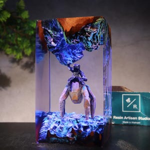 May include: A rectangular resin block featuring a miniature figure of a person riding a multi-legged creature. The scene includes blue and purple glowing textures resembling a landscape, with a wooden top section and a Resin Artisan Studio logo.