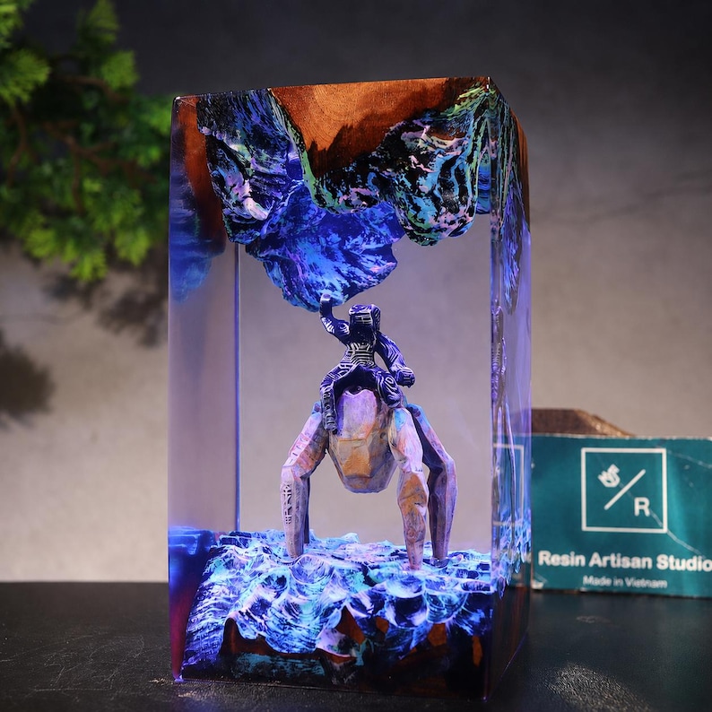 May include: A rectangular resin block featuring a miniature figure of a person riding a multi-legged creature. The scene includes blue and purple glowing textures resembling a landscape, with a wooden top section and a Resin Artisan Studio logo.