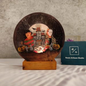 K&ouml;nnte beinhalten: Ein runder, transparenter Schneekugel aus Harz mit Halloween-Motiv. Die Kugel zeigt ein Spukhaus, einen gruseligen Clown und eine Figur mit Hockeymaske. Der Sockel ist aus Holz und hat ein Licht im Inneren.