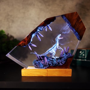 Puede incluir: Escultura decorativa de resina con una figura de dinosaurio azul y blanco. La escultura está encerrada en una forma geométrica transparente con detalles en madera y morado. Se apoya sobre una base de madera.