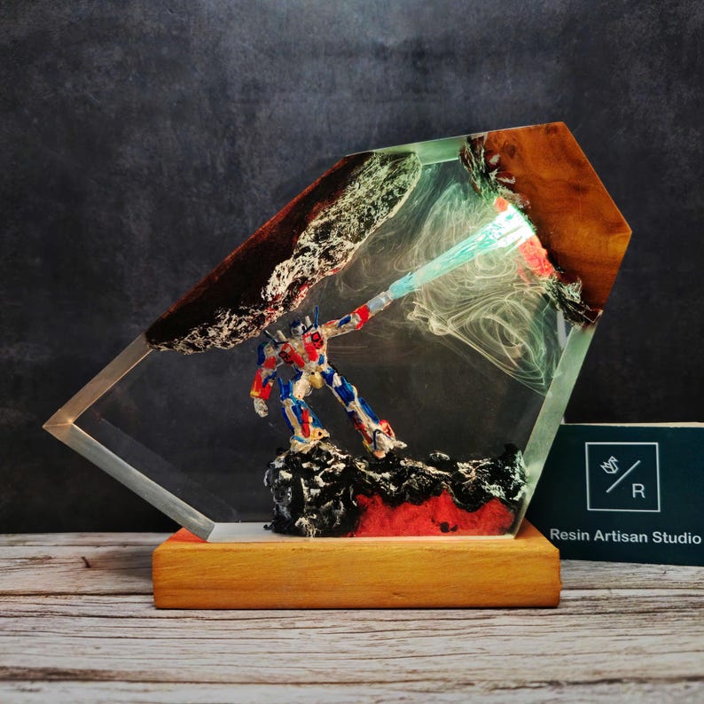 May include: A resin sculpture of a robot holding a glowing blue energy weapon, encased in a clear diamond-shaped block with a wooden base. The resin is swirled with black, red, and green colors, creating a dramatic effect. The sculpture is titled "Resin Artisan Studio".