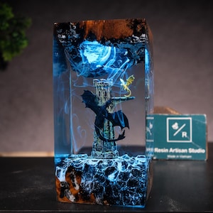 May include: A rectangular resin block lamp featuring a miniature stone tower and two dragons inside. A large black dragon is perched on the tower, while a smaller yellow dragon sits on a ledge. The interior glows with blue light and swirling smoke effects.