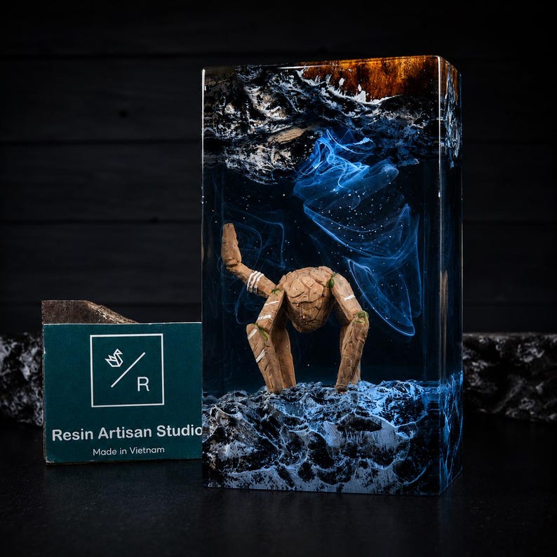 May include: A rectangular resin block featuring a submerged wooden figure with white markings, surrounded by swirling blue and white smoke-like patterns. The base is textured dark rock, and a green card with the text Resin Artisan Studio is visible.
