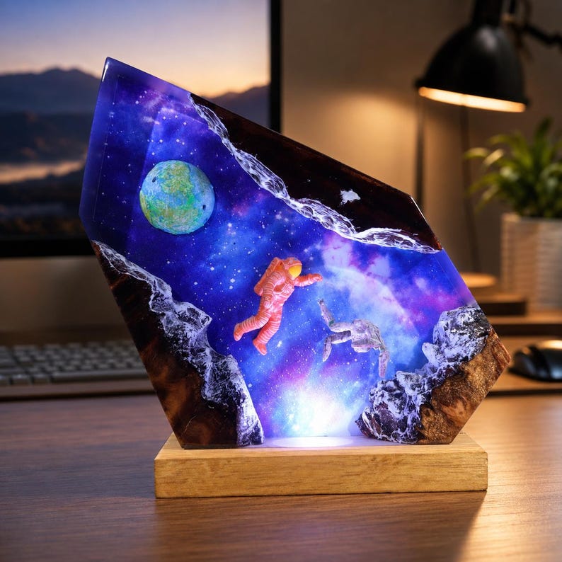 May include: A decorative resin lamp featuring a space scene with a blue and purple nebula, a green planet, and two floating astronaut figures. The resin is encased between two jagged pieces of dark wood and sits on a rectangular wooden base.