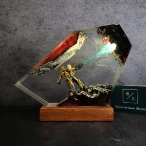 May include: A yellow robot figurine with a blue energy beam shooting out of its hand, encased in clear resin with a wooden base. The resin also contains red and white accents.