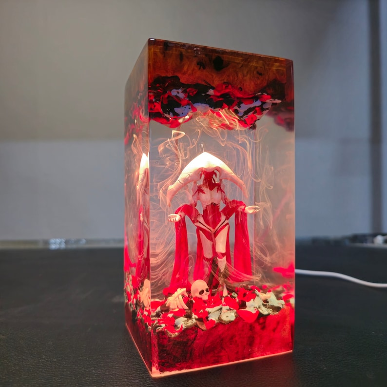 May include: A resin sculpture of a figure in a red dress and a white headpiece. The figure is standing on a bed of red and brown resin with a skull and bones. The sculpture is encased in a clear resin block.