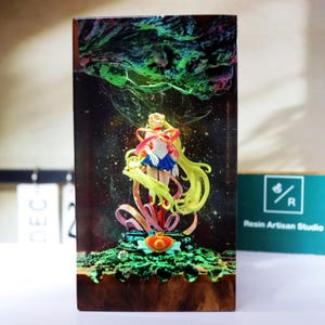 May include: A resin art piece featuring a Sailor Moon figurine. The figurine is in a dynamic pose, with long yellow hair and a colorful outfit. The background has a cosmic design with green and blue accents. The base is a detailed, textured surface.