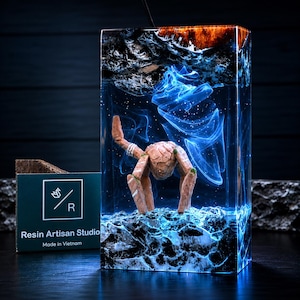 May include: A rectangular resin block lamp featuring a submerged tan crab-like creature. The interior displays swirling blue light effects, a dark textured top, and a rocky base. A green card with the text Resin Artisan Studio Made in Vietnam is visible.