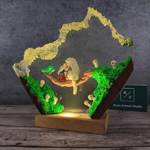 May include: A resin art lamp with a white wolf and small white figures in a forest scene. The lamp is lit and has a wooden base. The text "Resin Artisan Studio" is on a green card in the background.