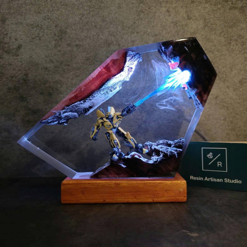 May include: A resin sculpture of a yellow robot holding a blue energy weapon, set in a clear resin block with a wood base. The sculpture is lit from behind, creating a glowing effect. The base has the text "Resin Artisan Studio" on it.