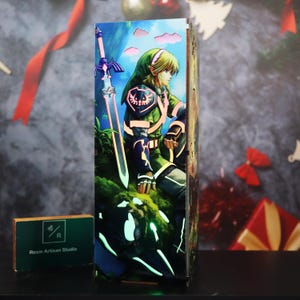 Puede incluir: Una pieza de arte de resina rectangular e iluminada que presenta un personaje de The Legend of Zelda. La obra de arte representa una figura vestida de verde con una espada sobre un fondo de cielo azul. La pieza tiene un efecto brillante.