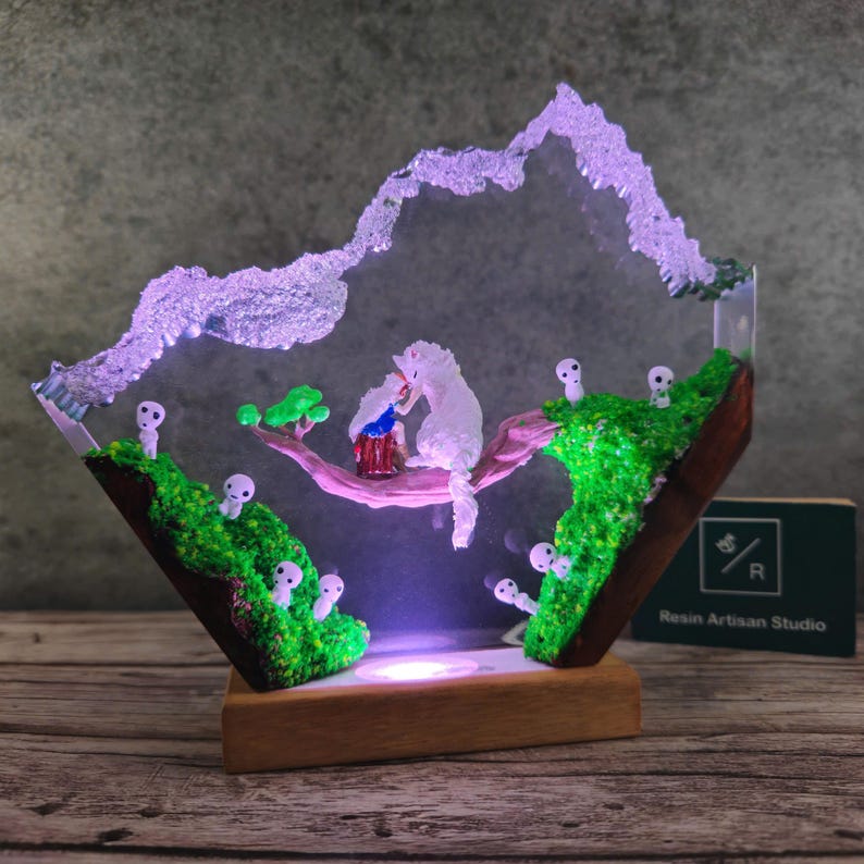 May include: A resin art piece with a white and blue figurine of a character from the movie Spirited Away, sitting on a branch with a green tree and white figures on a green mossy base. The piece is lit from below with a white light and has a wooden base with the text "Resin Artisan Studio".