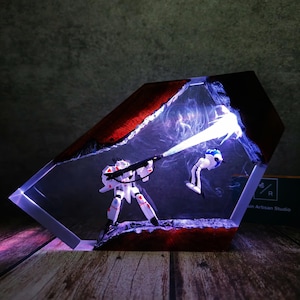 Puede incluir: Diorama futurista con dos figuras de robots en una caja hexagonal de resina. Un robot blanco y rojo dispara un rayo a otro robot que cae. La caja tiene un borde rojo y está iluminada con luces azules y moradas.