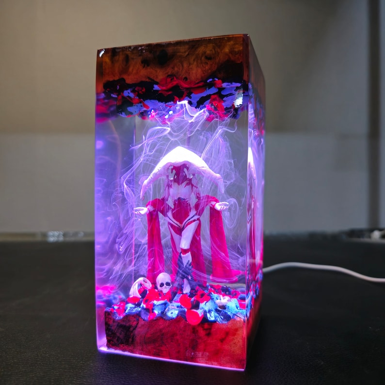 May include: A clear resin cube with a red and white figure inside. The figure is wearing a white hooded cloak and a red dress. The cube is lit from the inside, creating a purple glow around the figure. There are red and blue glitter and a skull at the bottom of the cube.