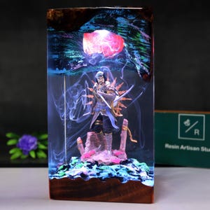 Puede incluir: Una pieza de arte de resina rectangular con una figura de guerrero con una espada, sobre un fondo de un orbe rojo y patrones arremolinados azules y verdes. La base y la parte superior son de madera oscura. La pieza está iluminada con iluminación interna.