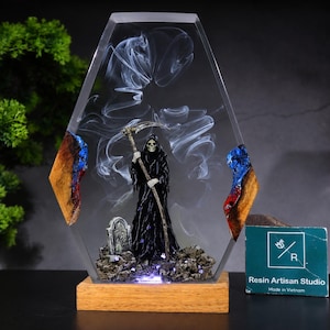 May include: A coffin-shaped resin display featuring a grim reaper figure in a black robe holding a scythe, standing on a rocky base with a tombstone. The clear resin contains swirling white smoke effects and wood accents. Includes a wooden base.