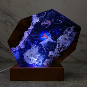 May include: A hexagonal resin lamp with a blue and white galaxy scene, featuring a white spaceship and red fish. The lamp is lit with blue LED lights and sits on a wooden base.