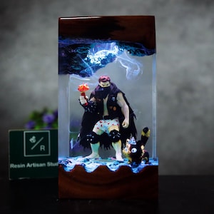 May include: A resin art piece featuring a muscular figure in a black cloak and shorts with heart patterns, holding a torch. A black cat-like figure stands nearby. The artwork is encased in a clear resin block with blue and white accents, and a wooden base.