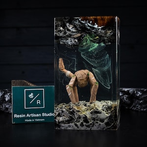 May include: A rectangular resin block featuring a miniature wooden creature with four legs standing on a textured base. The clear resin contains swirling green and white smoke-like patterns, with dark wood elements at the top and bottom edges.